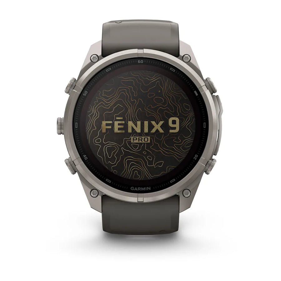A Garmin Fenix 9 Pro smartwatch with a round silver-toned metal case, dark gray strap, and a black display showing a topographic map pattern with gold lettering.